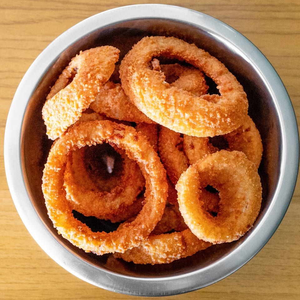 A metal bowl filled with golden-brown, crispy onion rings from The Cheesesteak Grill.