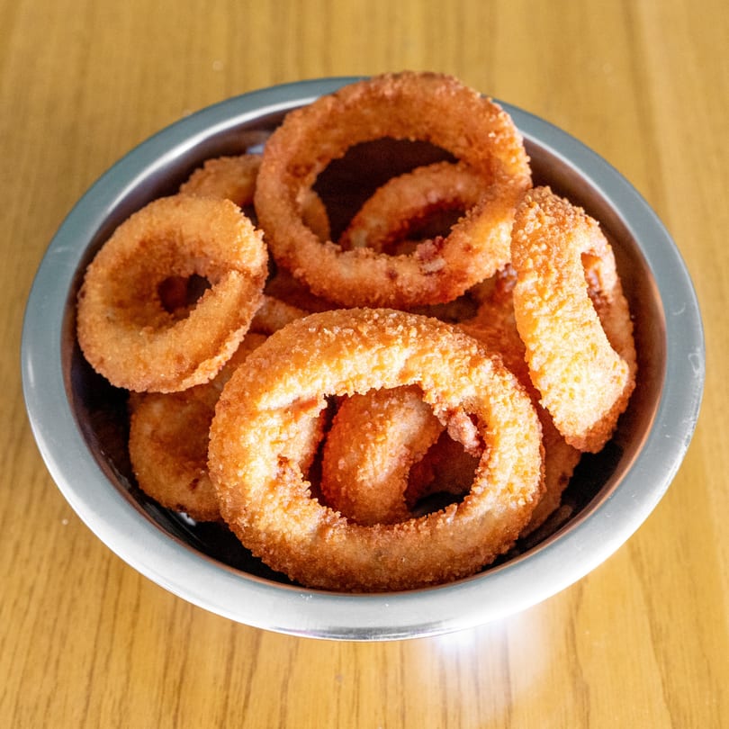 A metal bowl filled with golden-brown, crispy onion rings on a light wooden table.